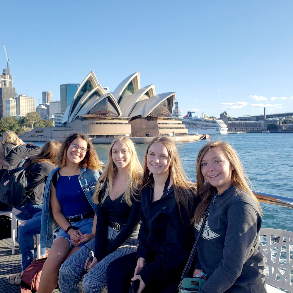 four girls sitting at the dock of the bay with the sydney opera house in the background
